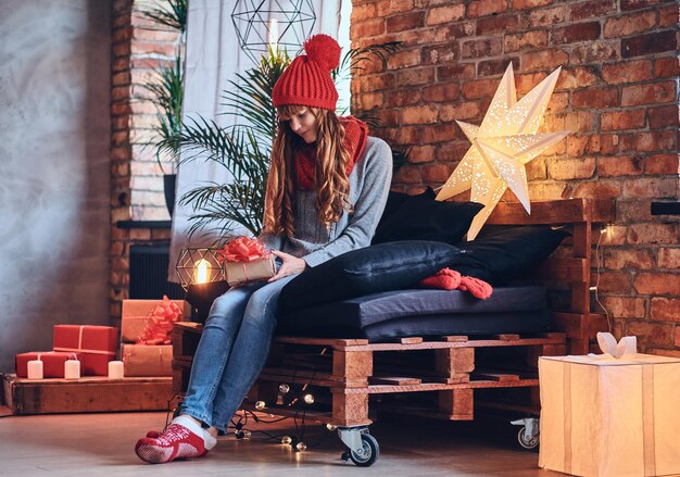 Rothaarige Frau hält ein Weihnachtsgeschenk in einem Wohnzimmer mit Loft-Interieur.