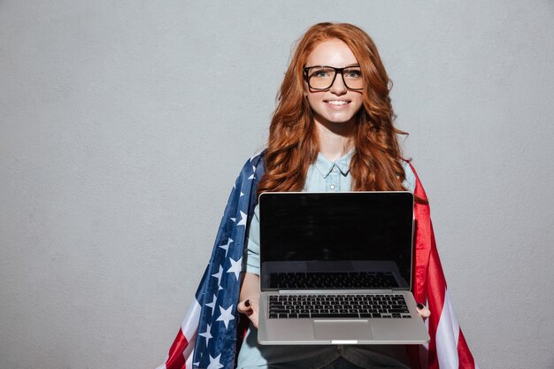 Rothaarige junge Dame mit USA-Flagge, die Anzeige des Laptops zeigt