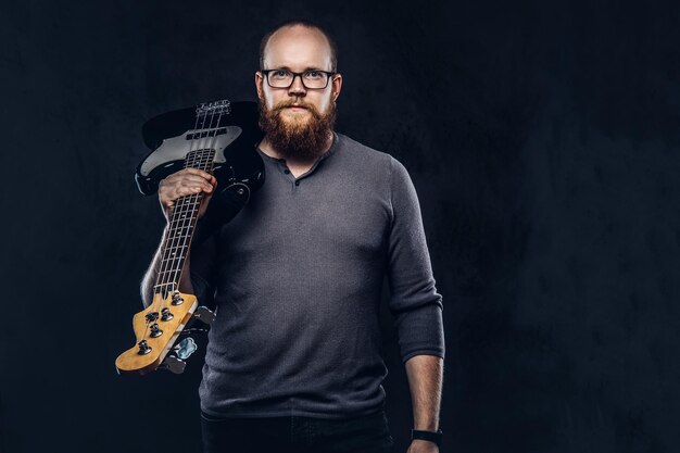 Rothaariger bärtiger männlicher Musiker mit Brille in einem grauen T-Shirt hält E-Gitarre. Getrennt auf dunklem strukturiertem Hintergrund.