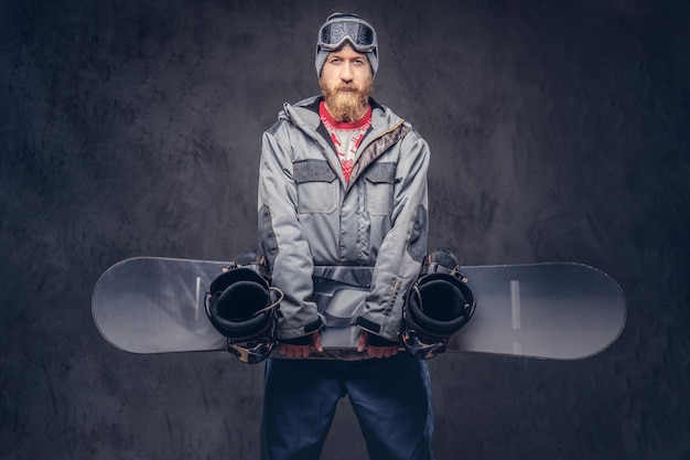 Kostenloses Foto rothaariger bärtiger snowboarder mit wintermütze und schutzbrille in einem snowboardmantel hält ein snowboard in einem studio. getrennt auf einem dunklen strukturierten hintergrund.
