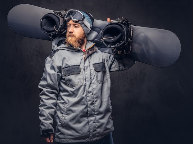 Kostenloses Foto rothaariger bärtiger snowboarder mit wintermütze und schutzbrille in einem snowboardmantel posiert mit snowboard in einem studio und schaut weg. isoliert auf grauem hintergrund.