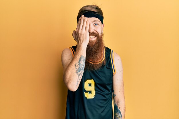 Rothaariger Mann mit langem Bart in Basketballuniform, der ein Auge mit der Hand bedeckt, selbstbewusstes Lächeln im Gesicht und überraschende Emotionen.