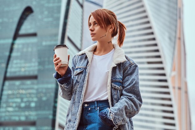 Rothaariges Hipster-Mädchen mit Tätowierung auf ihrem Gesicht, das Jeansmantel trägt und Kaffee zum Mitnehmen vor Wolkenkratzern in der Stadt Moskau hält.