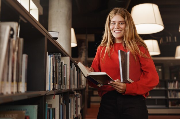 rothaariges Mädchen, Student, der in der Bibliothek nahe Regalen steht, ein Buch liest und lächelt.