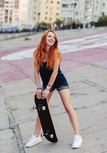 Rothaariges, schlankes Mädchen in Jeansshorts und schwarzem T-Shirt posiert mit Skateboard in urbaner Sciene.