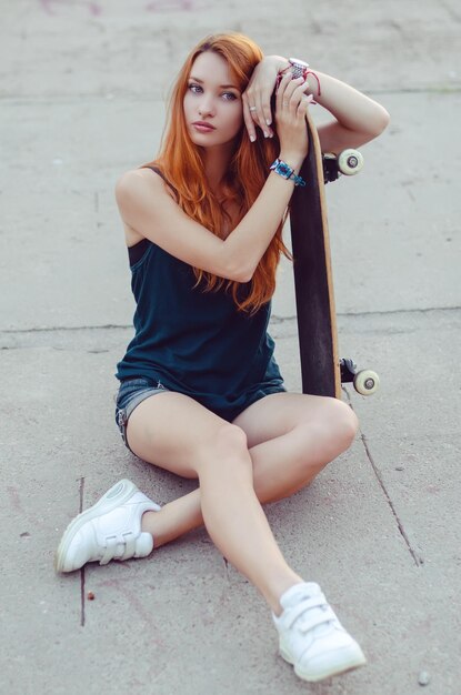 Rothaariges, schlankes Mädchen in Jeansshorts und schwarzem T-Shirt posiert mit Skateboard in urbaner Sciene.