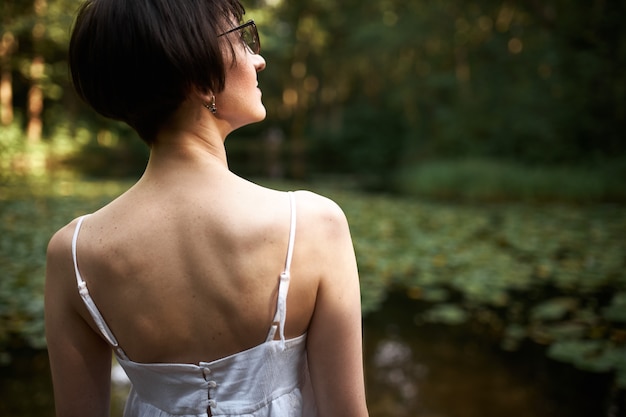 Rückansicht der attraktiven dunkelhaarigen Frau mit dem kurzen Haarschnitt, der im Wald posiert, der Brillen und weißes Riemenkleid trägt, das am Teich steht und ruhige friedliche Atmosphäre genießt.