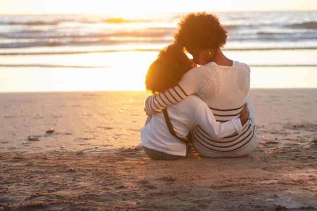 Rückansicht der liebevollen Mutter und Tochter, die den Sonnenuntergang betrachten. Mutter und Tochter sitzen auf Sand, umarmen sich und sehen sich an. Familie, Liebe, Bindung, Naturkonzept