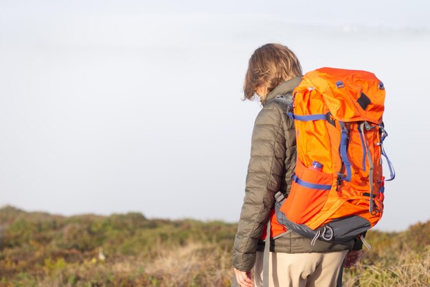 Rückansicht der Wanderin am Sommertag. Frau mit großem orangefarbenem Rucksack, der auf der Wiese steht. Hobby, aktives Lifestyle-Konzept