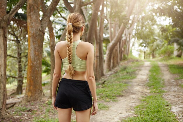 Rückansicht des Joggers der jungen Frau mit fitem Körper, der Sport-BH und schwarze Shorts trägt, die allein auf Spur im tropischen Wald stehen, der bestimmt ist, zu laufen.