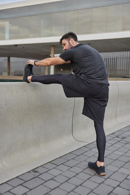 Rückansicht des männlichen Joggers macht Dehnübungen trainiert Muskeln bereitet sich auf das Training im Freien vor genießt einen gesunden Lebensstil hat Training in der Stadtstraße trägt schwarze Sportkleidung erreicht Fitnessziele