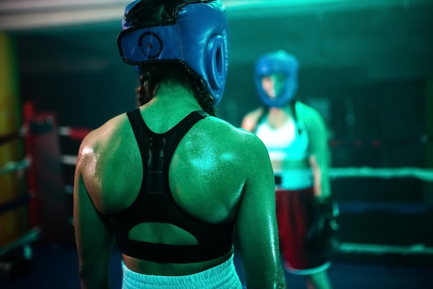 Rückansicht des starken Rückens der Boxermädchen während des aktiven Kampfes. Zwei junge Mädchen in Helmen stehen sich gegenüber und schauen sich an, um sich auf ein neues Sparring am Ring vorzubereiten. Gesunder Lebensstil, Extremsportkonzept