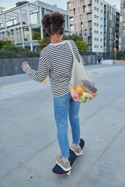 Rückansicht einer aktiven Frau mit lockigem Haar genießt Skateboarding in der Stadt, die an Extremsportarten beteiligt ist, kehrt vom Markt mit einer Netztasche mit Gemüse zurück und hört Lieblingslied in Kopfhörern Urbaner Lebensstil