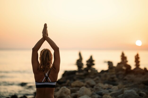 Rückansicht einer Frau, die bei Sonnenuntergang Yoga-Entspannungsübungen am Meer macht. Platz kopieren.