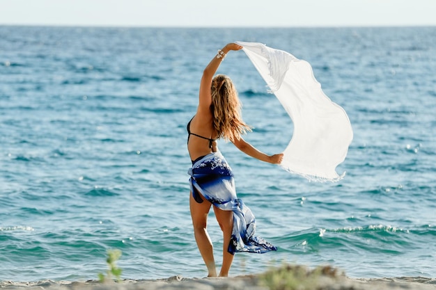 Rückansicht einer Frau, die sich frei fühlt, während sie an einem windigen Tag am Meer einen Schal hält