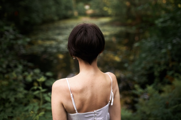 Rückansicht einer nicht wiedererkennbaren jungen Frau mit kurzen Haaren und schlankem Körper, die draußen posiert, vor Teich mit ihrem Rücken zur Kamera stehend.