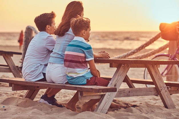Rückansicht einer süßen Familie - Mutter mit ihren Söhnen sitzt auf einer Bank und blickt auf den Sonnenuntergang an der Küste.