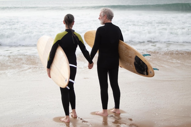 Rückansicht eines älteren Paares, das einen Spaziergang mit Surfbrettern genießt. Sportlicher grauhaariger Mann und Frau, die zusammen am Meer spazieren gehen und sich an den Händen halten. Aktive Erholung, Freizeitbeschäftigung des Alterskonzepts