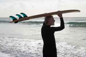 Kostenloses Foto rückansicht eines alten mannes mit surfbrett. bärtiger kaukasischer mann, der am ufer steht und ein brett auf dem kopf hält und die schöne aussicht auf das meer und die wellen genießt. sportliche aktivität, aktive erholung für ältere menschen