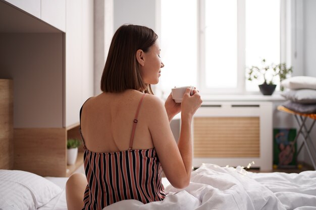 Rückansicht Frauen, die Kaffee auf dem Bett genießen, während sie sich nach dem Aufwachen strecken.