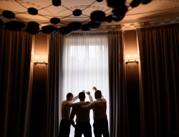 Rückansicht Freunde von Bräutigammännern heben Gläser mit Getränken, die vor einem großen Fenster in einer luxuriösen, gemütlichen Wohnung stehen und Junggesellenabschied feiern und auf den Hochzeitstag warten