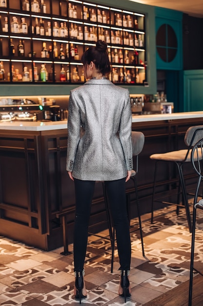 Rückansicht in voller Länge des Modemodells in funkelnder silberner Jacke, schwarzer Hose und High Heels mit Frisur in trendiger Bar.