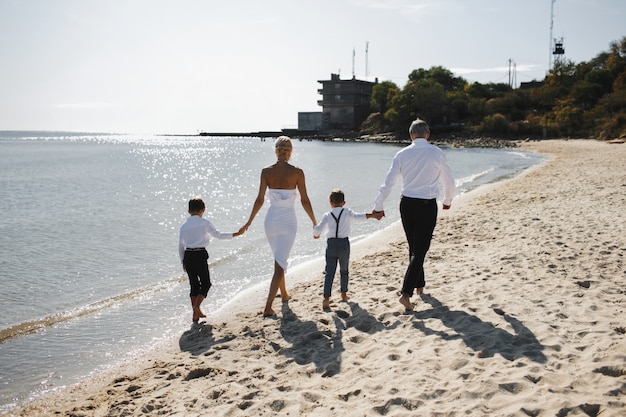 Rückansicht von Eltern und Kindern halten Hände zusammen und gehen am sonnigen Sommertag am Strand spazieren, gekleidet in weiße stilvolle Kleidung