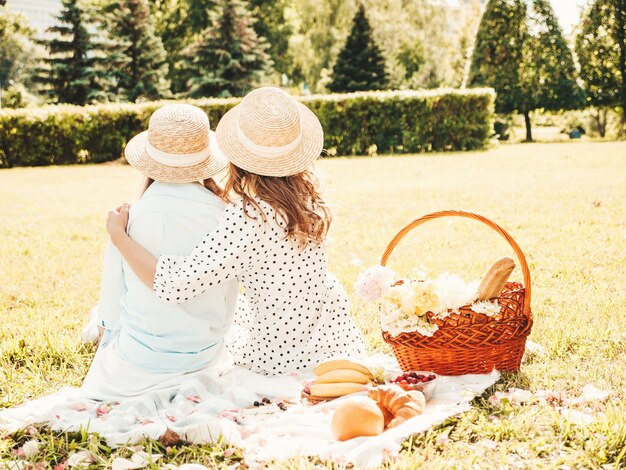 Rückansicht von zwei jungen schönen lächelnden Hippie-Mädchen in trendigem Sommerkleid und Hüten. Frauen, die draußen Picknick machen.