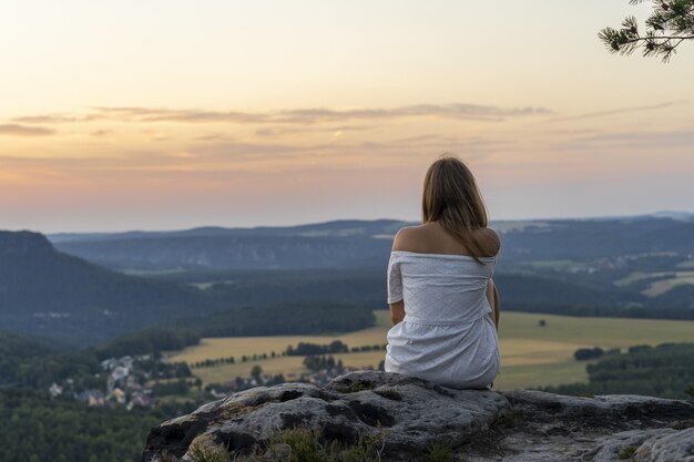 Rückansichtaufnahme einer jungen Frau, die am Rand einer Klippe sitzt und einen majestätischen Sonnenuntergang genießt