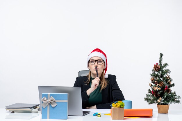 Rücksichtsvolle blonde Frau mit einem Weihnachtsmannhut, der an einem Tisch mit einem Weihnachtsbaum und einem Geschenk auf ihm auf weißem Hintergrund sitzt