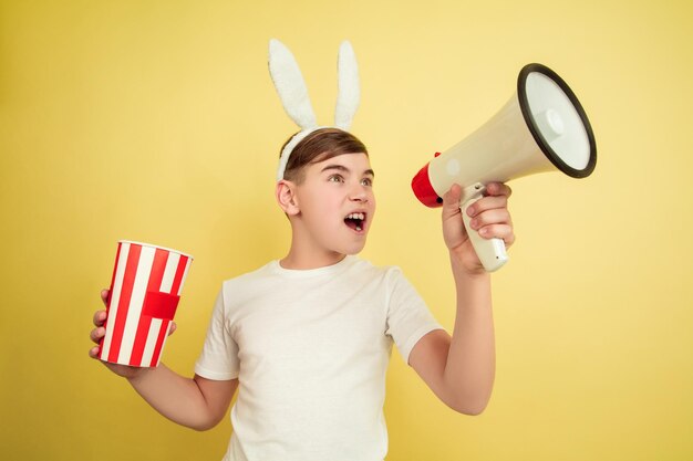 Rufen mit Trompete, Popcorn. Kaukasischer Junge als Osterhase auf gelbem Hintergrund. Fröhliche Ostergrüße.