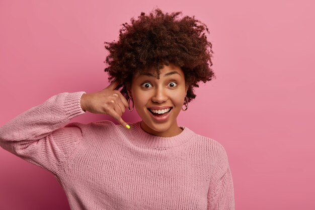 Rufen Sie mich irgendwann an. Eine zufriedene ethnische Frau mit Afro-Haaren macht eine Telefongeste, bittet die Person um Kontaktnummer, lächelt positiv, schlägt vor, dass Sie hängen, trägt einen rosigen Pullover.