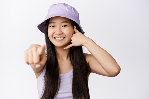 Rufen Sie uns an. Porträt des brünetten asiatischen Mädchens im Eimerhut, das Telefonanrufgeste zeigt und Finger auf Weiß zeigt, glücklich auf Weiß lächelt.