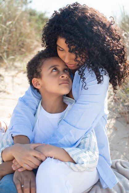 Ruhige afroamerikanische Mutter und Kind am Strand. Mutter und Sohn in Freizeitkleidung sitzen auf einer Decke und umarmen sich. Familie, Entspannung, Naturkonzept