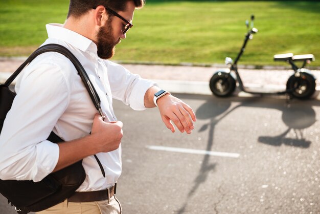 Ruhiger bärtiger Mann in der Sonnenbrille, die Rucksack hält und auf seine Armbanduhr schaut, während im Freien mit modernem Motorrad auf Hintergrund aufwirft