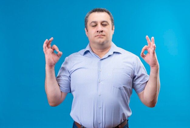Ruhiger Mann mittleren Alters im blau gestreiften Hemd, der die Augen geschlossen hält und eine gute Geste mit den Händen auf einem blauen Raum macht
