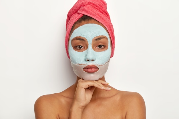 Ruhiges junges weibliches Model genießt tägliche Hautpflege, trägt eine beruhigende Maske auf das Gesicht auf, reduziert den Oberflächenglanz, peelt Mitesser, hat Handtuch auf den Kopf gewickelt, berührt das Kinn sanft und sieht aus