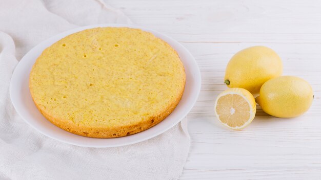 Runder Zitronenkuchen diente in der weißen Platte mit ganzen Zitronen auf hölzernem Hintergrund