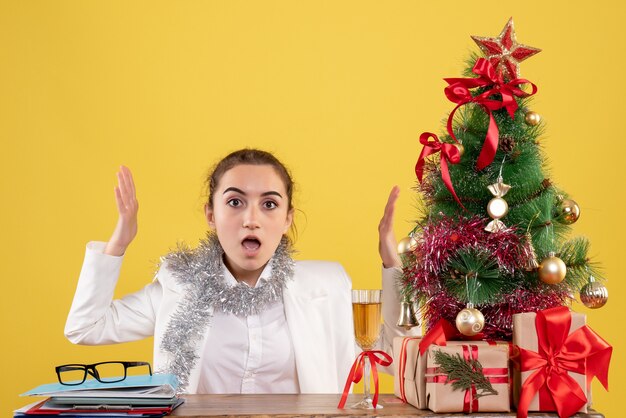 Ärztin der Vorderansicht, die hinter ihrem Tisch auf gelbem Hintergrund mit Weihnachtsbaum und Geschenkboxen sitzt