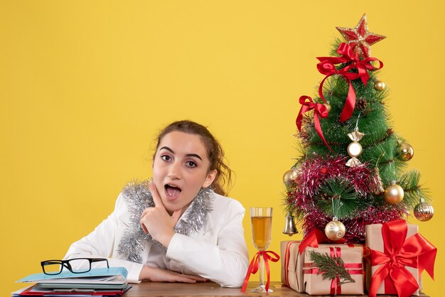 Ärztin der Vorderansicht, die hinter ihrem Tisch auf gelbem Hintergrund mit Weihnachtsbaum und Geschenkboxen sitzt