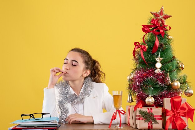 Ärztin der Vorderansicht, die hinter ihrem Tisch auf gelbem Hintergrund mit Weihnachtsbaum und Geschenkboxen sitzt