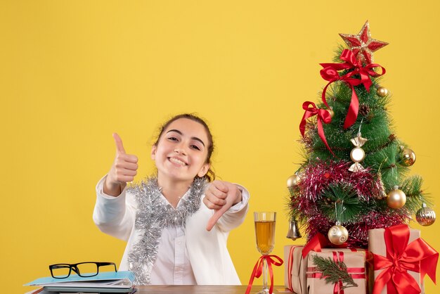 Ärztin der Vorderansicht, die hinter ihrem Tisch auf gelbem Hintergrund mit Weihnachtsbaum und Geschenkboxen sitzt