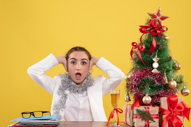 Ärztin der Vorderansicht, die hinter ihrem Tisch auf gelbem Hintergrund mit Weihnachtsbaum und Geschenkboxen sitzt