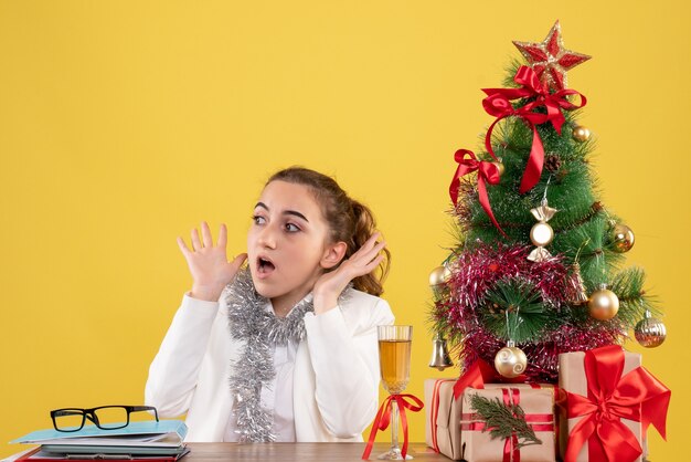 Ärztin der Vorderansicht, die hinter ihrem Tisch mit verängstigtem Gesicht auf gelbem Hintergrund mit Weihnachtsbaum und Geschenkboxen sitzt