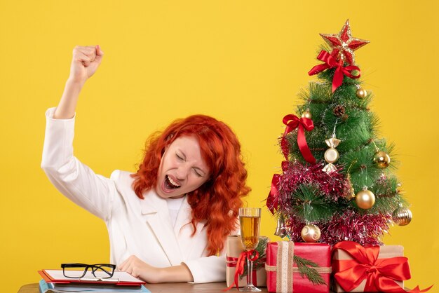 Ärztin der Vorderansicht, die hinter ihrem Tisch mit Weihnachtsgeschenken und Baum auf gelbem Hintergrund sitzt