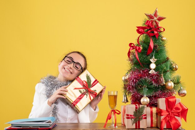 Ärztin der Vorderansicht, die hinter ihrem Tisch mit Weihnachtsgeschenken und Baum auf gelbem Hintergrund sitzt
