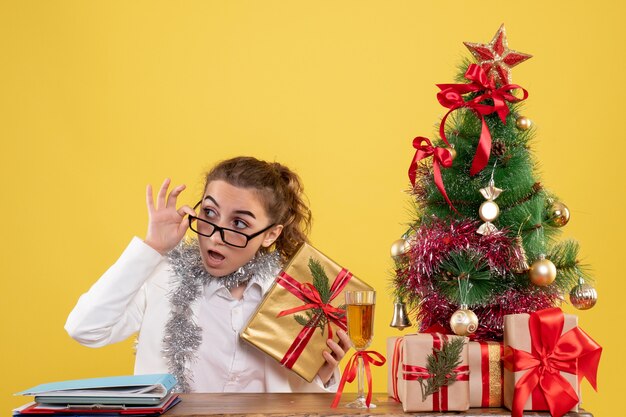 Ärztin der Vorderansicht, die hinter ihrem Tisch mit Weihnachtsgeschenken und Baum auf gelbem Hintergrund sitzt
