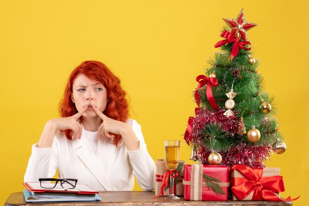 Ärztin der Vorderansicht, die hinter ihrem Tisch mit Weihnachtsgeschenken und Baum denkend auf gelbem Hintergrund sitzt