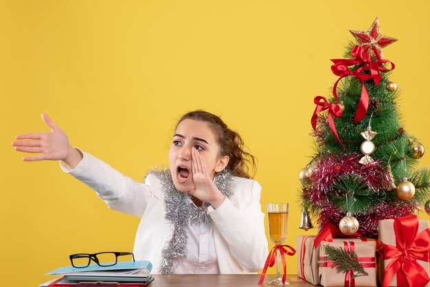 Ärztin der Vorderansicht, die hinter ihrem Tisch schreit, schreit auf gelbem Hintergrund mit Weihnachtsbaum und Geschenkboxen