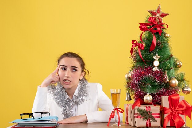 Ärztin der Vorderansicht, die hinter ihrem Tisch sitzt, betont auf gelbem Hintergrund mit Weihnachtsbaum und Geschenkboxen